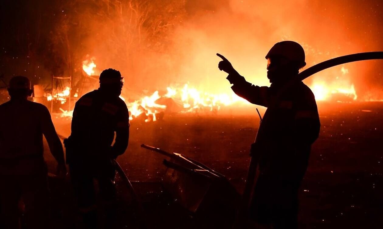 Πυροσβέστες επιχειρούν τη νύχτα μπροστά σε μεγάλη πυρκαγιά, με τις φλόγες να κατακαίνε εγκατάσταση και τον ουρανό να φωτίζεται από το πύρινο μέτωπο.